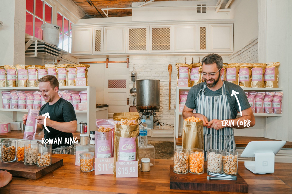 Two men in a commercial kitchen setting, surrounded by bags of popcorn and jars of corn kernels. One man on the left is labeled 'ROWAN RING' and is holding a pink bag of popcorn.